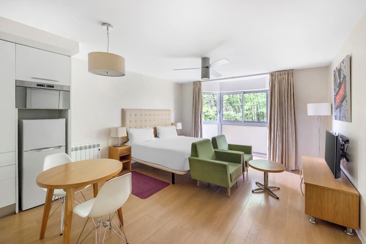 A modern hotel room with a bed, a desk, a chair, a small table, a TV, and a window with curtains.