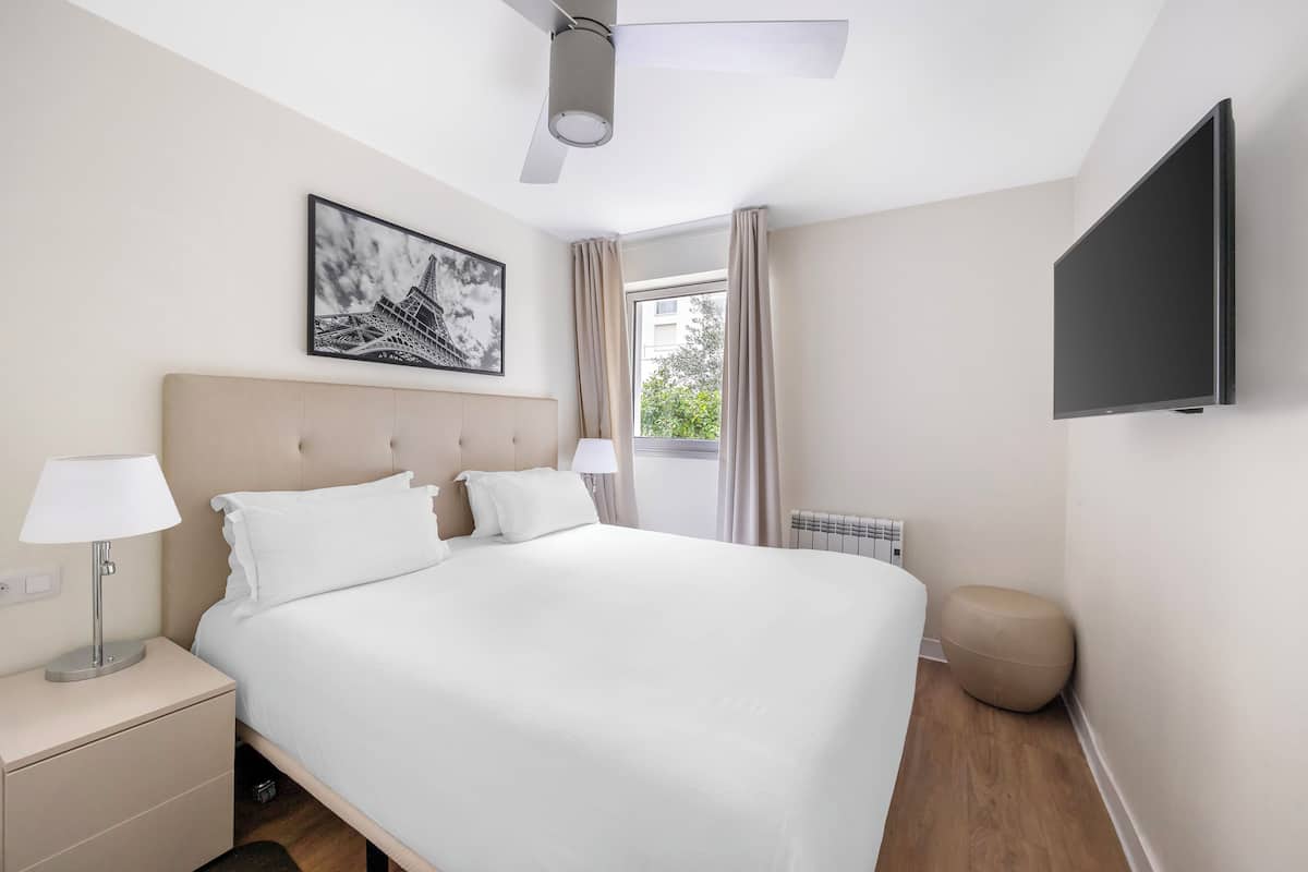 A hotel room with a bed, a nightstand, a lamp, a ceiling fan, a TV, and a window with curtains.