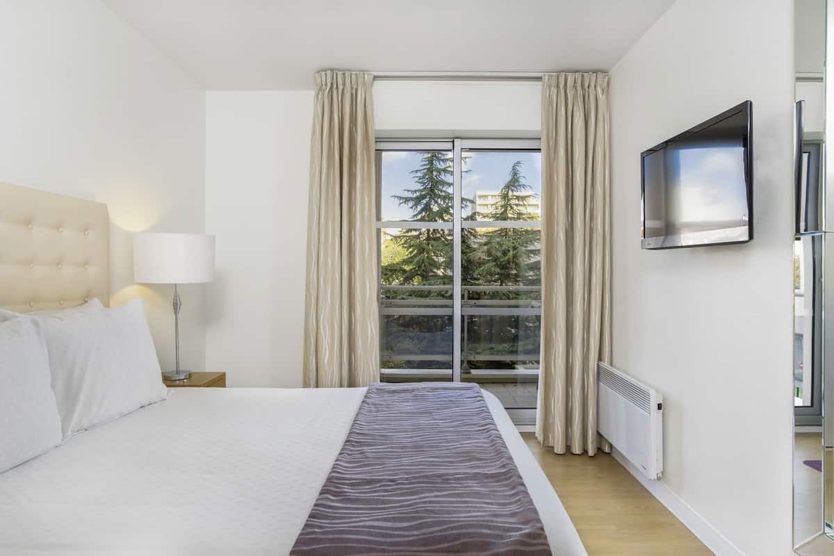 A hotel room with a bed, a TV, a window with curtains, and a view of trees and buildings.