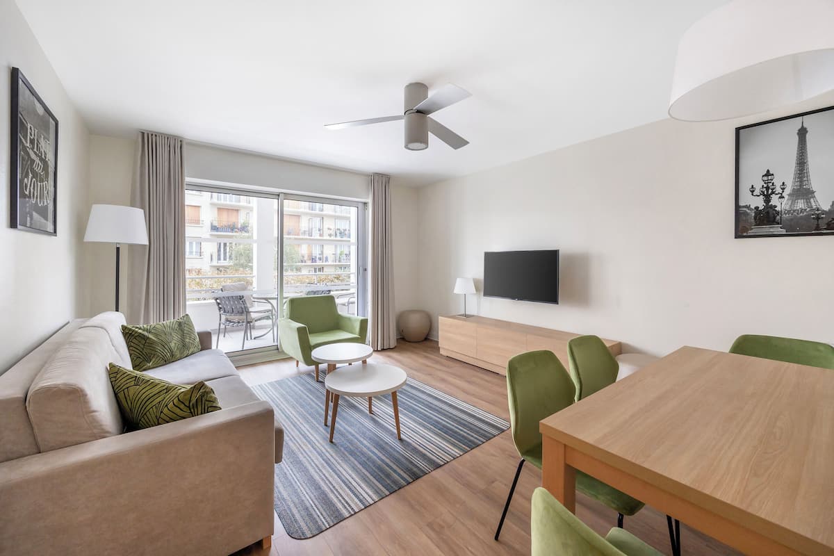A modern living room with a sofa, green chairs, a dining table, a ceiling fan, a TV, and a balcony with a view of buildings.