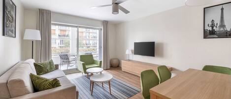 Een moderne woonkamer met een bank, groene stoelen, een eettafel, een plafondventilator, een televisie en een balkon met uitzicht op gebouwen.