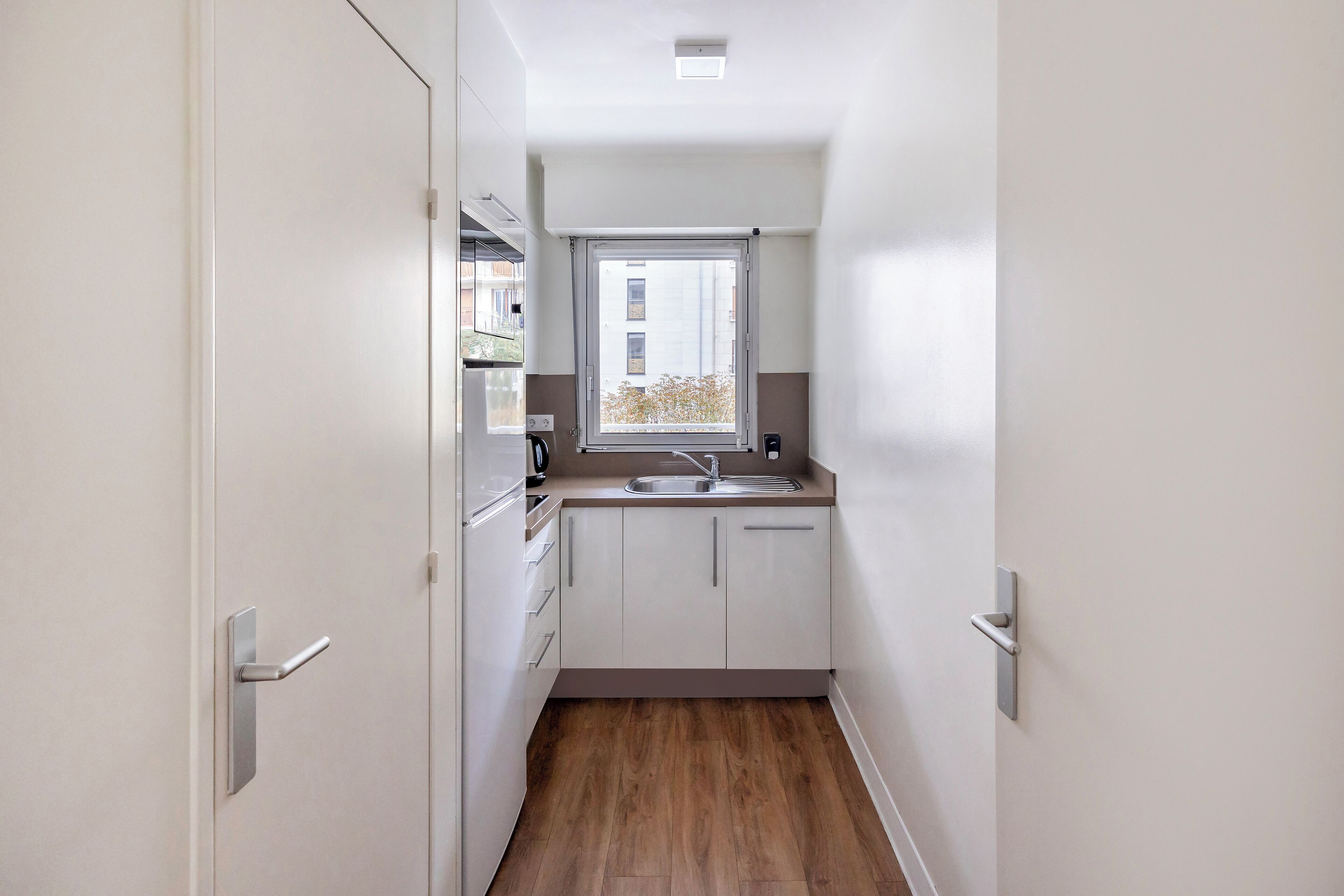 A narrow kitchen with white cabinets, a window, and a wooden floor.