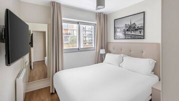 A hotel room with a large bed, a flat-screen TV, a window with curtains, and a cityscape painting on the wall.