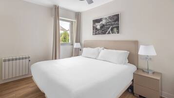 Un grand lit avec du linge de lit blanc, une table de chevet avec une lampe, un radiateur et une fenêtre avec des rideaux.