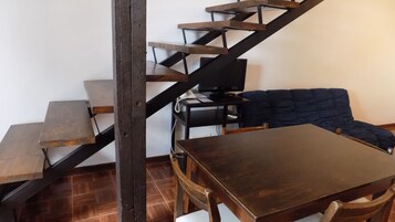 Una escalera con peldaños de madera y barandillas metálicas, una mesa de madera oscura, un sofá azul y un escritorio pequeño con una computadora y un teléfono.