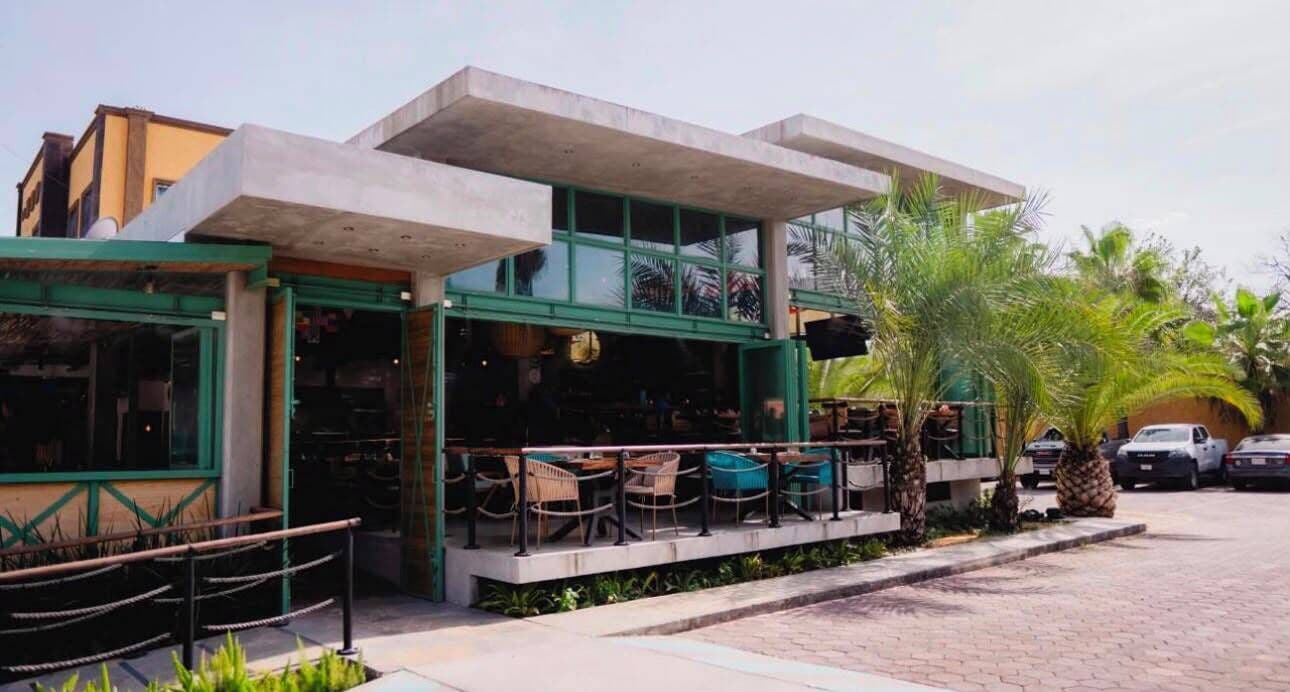 Un restaurante con mesas al aire libre, estructura de concreto y palmeras.