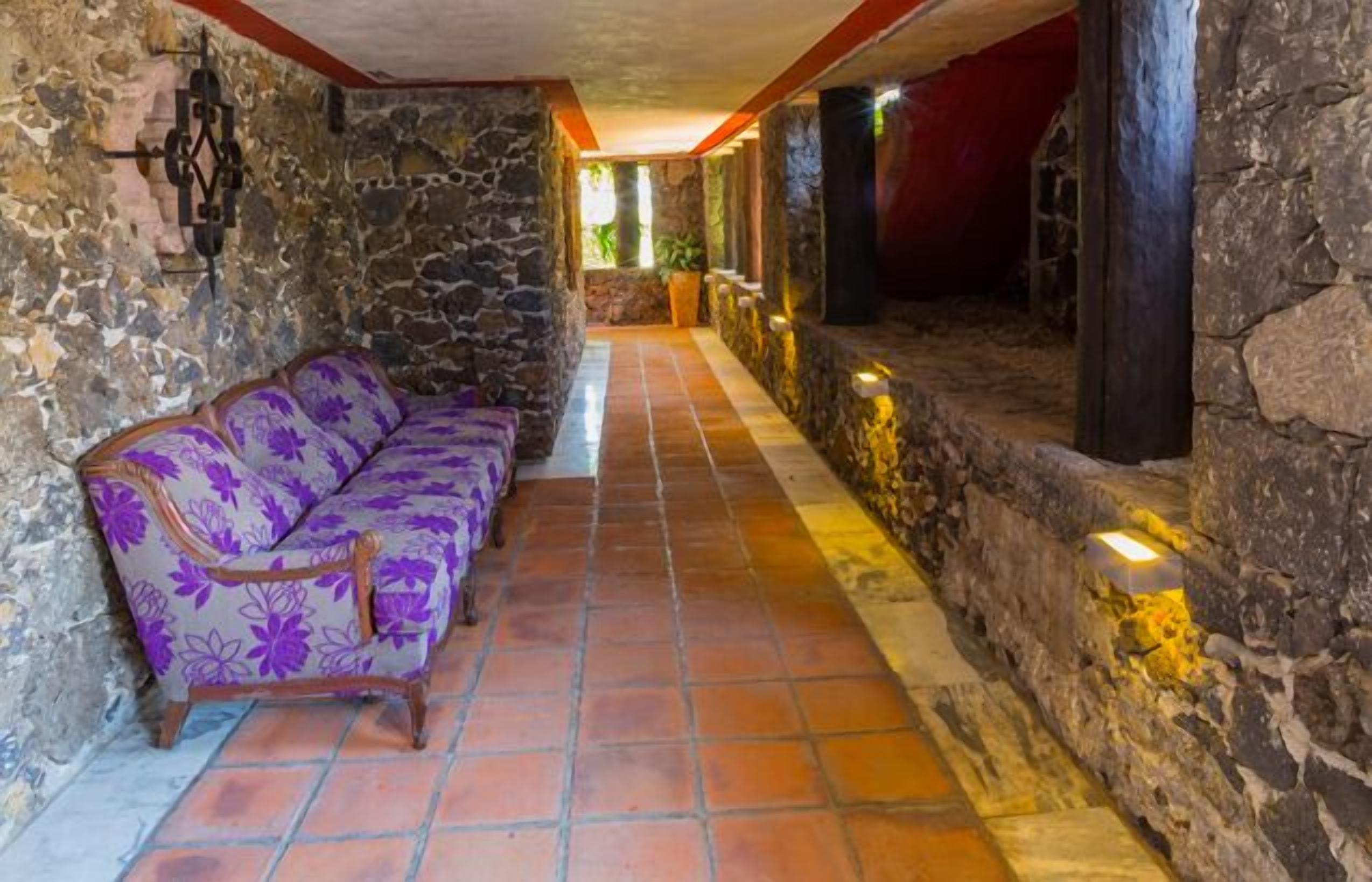A corridor with a stone wall, a purple floral sofa, and a tiled floor.