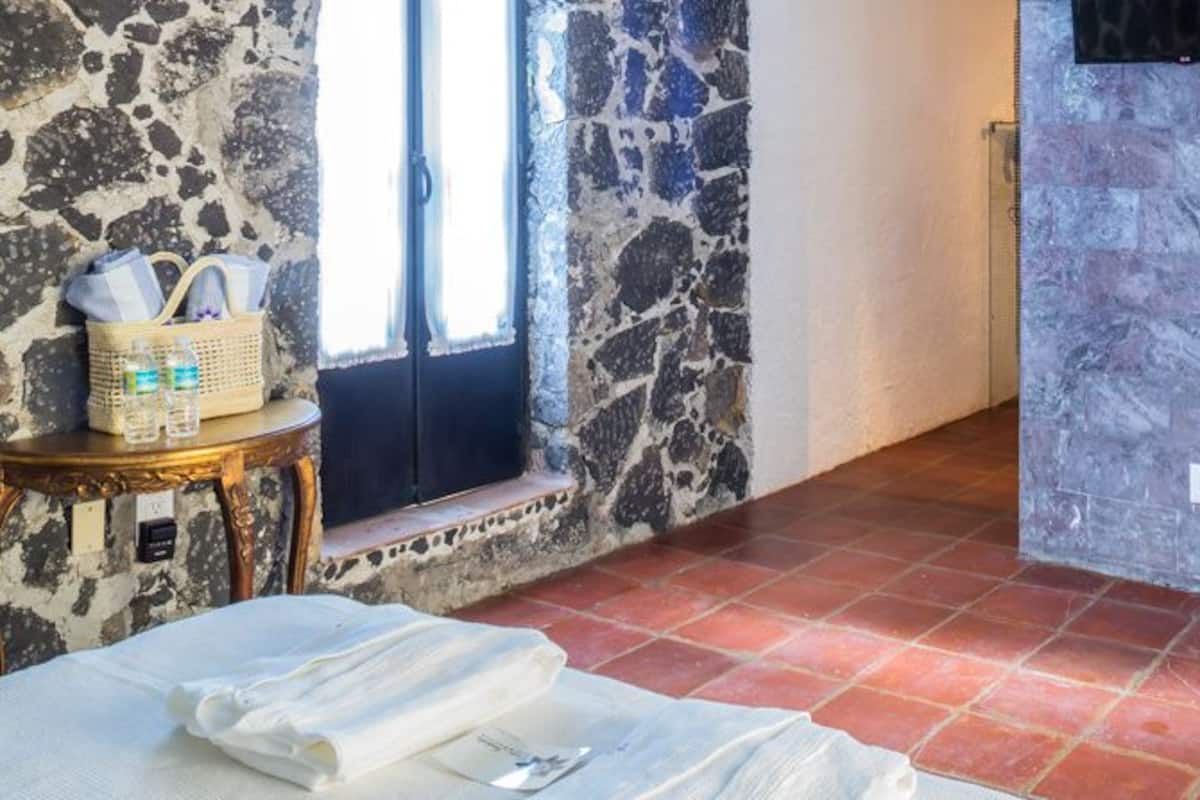 A room with a stone wall, a door, a bed with white linens, a small table with a basket and bottles, and a chair with a decorative cushion.