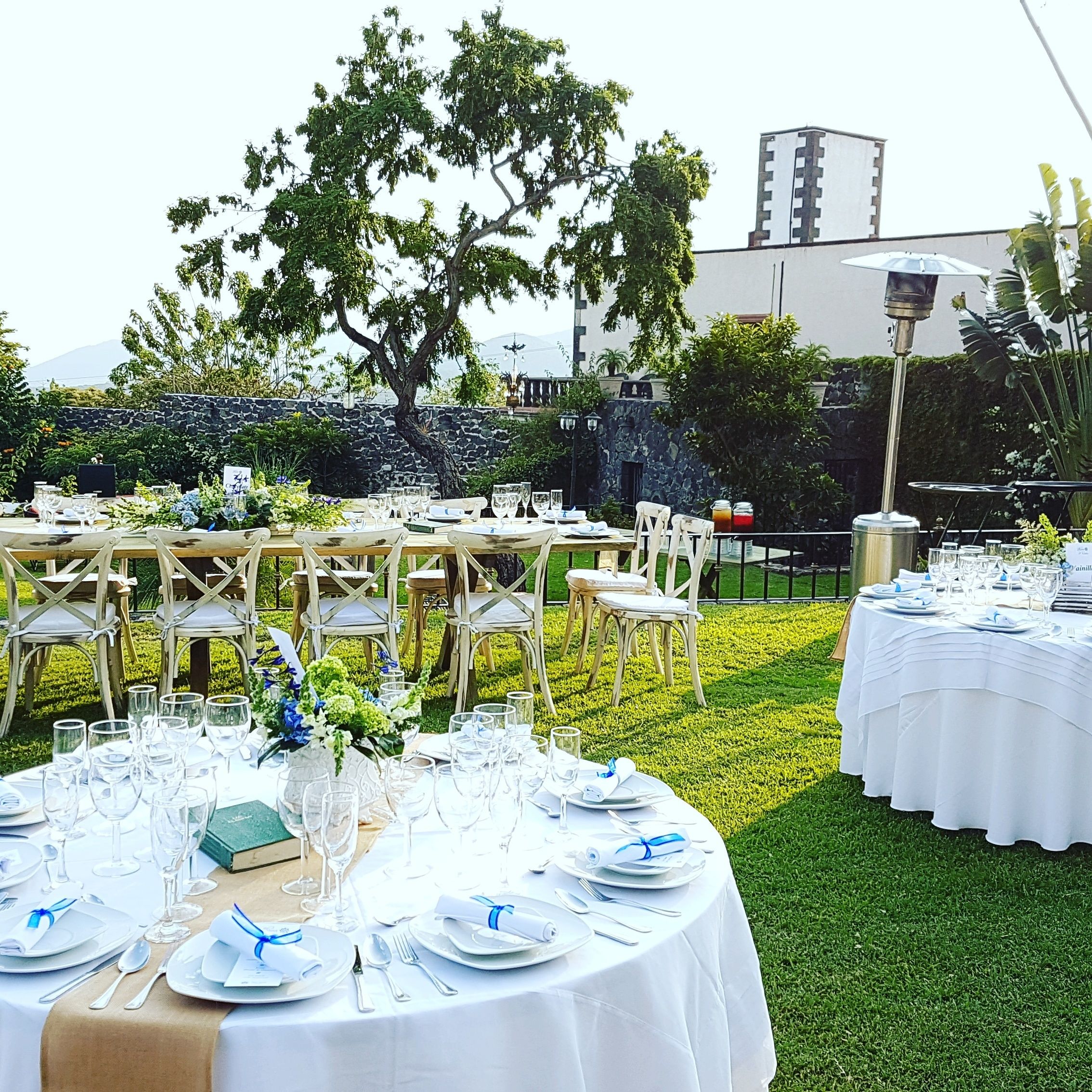 Un área de comedor al aire libre con mesas redondas dispuestas para un evento formal, rodeada de follaje y un muro de piedra.