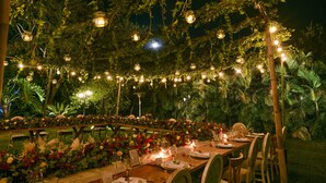 Un espace de restauration en plein air, avec des tables dressées pour le repas, entouré d’une végétation luxuriante et illuminé de guirlandes lumineuses.