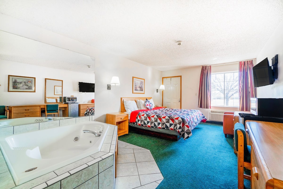 A hotel room with a large bathtub, a bed with a colorful geometric bedspread, a desk with a chair, and a television mounted on the wall.