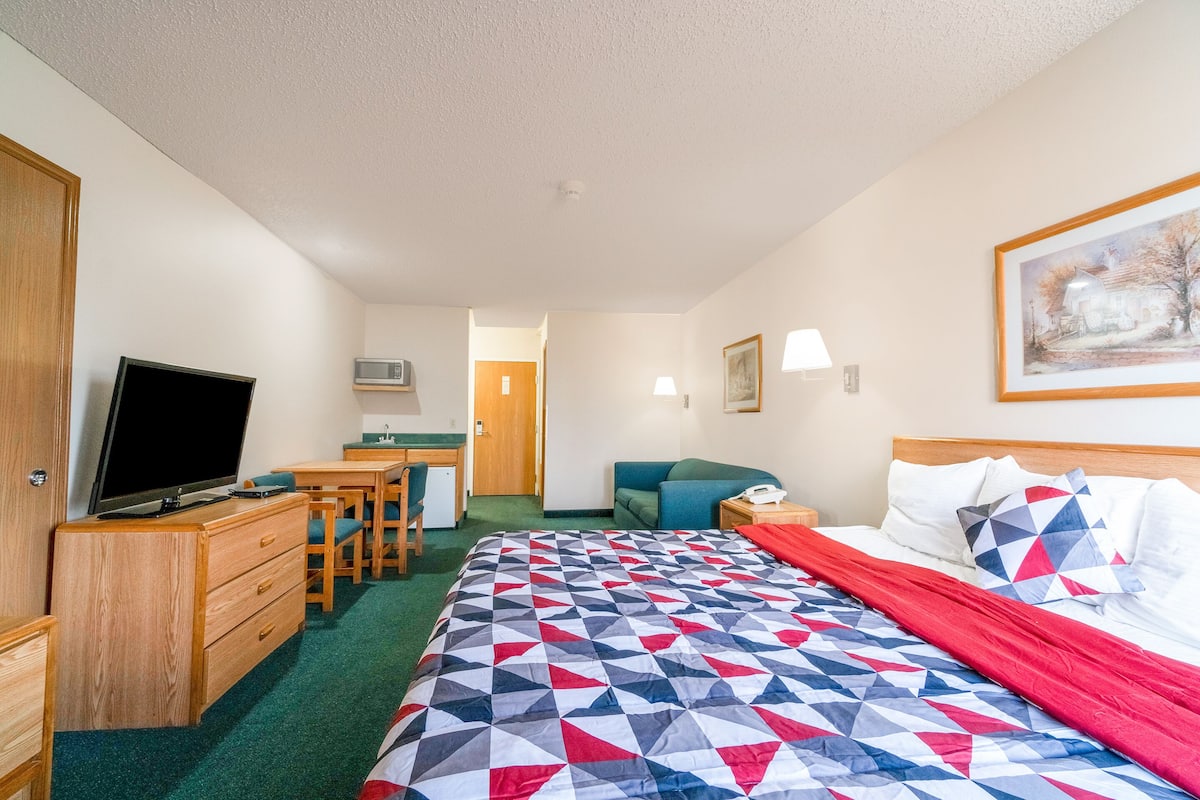 A hotel room with a bed, a desk with a chair, a television, a microwave, a couch, and a painting on the wall.