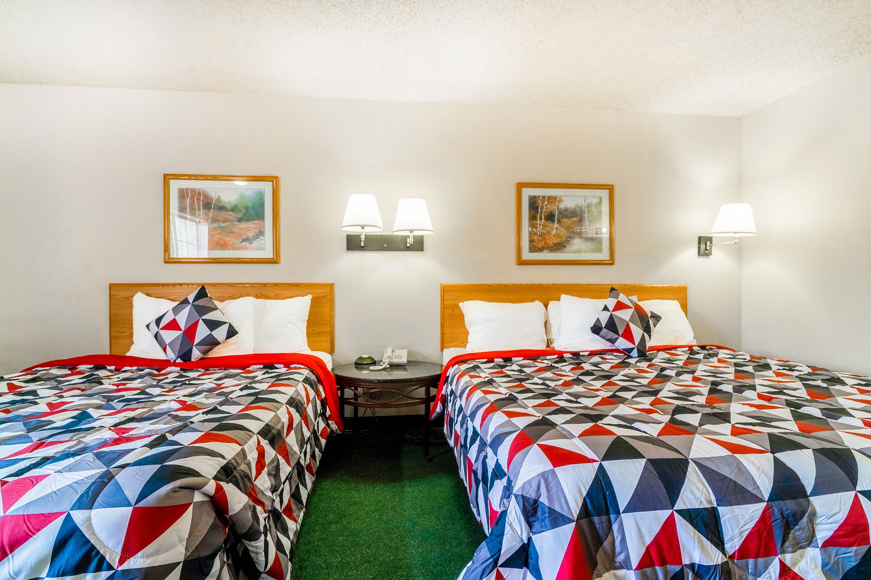 A hotel room with two beds, each with a geometric-patterned bedspread, a wooden headboard, and a nightstand with a lamp.