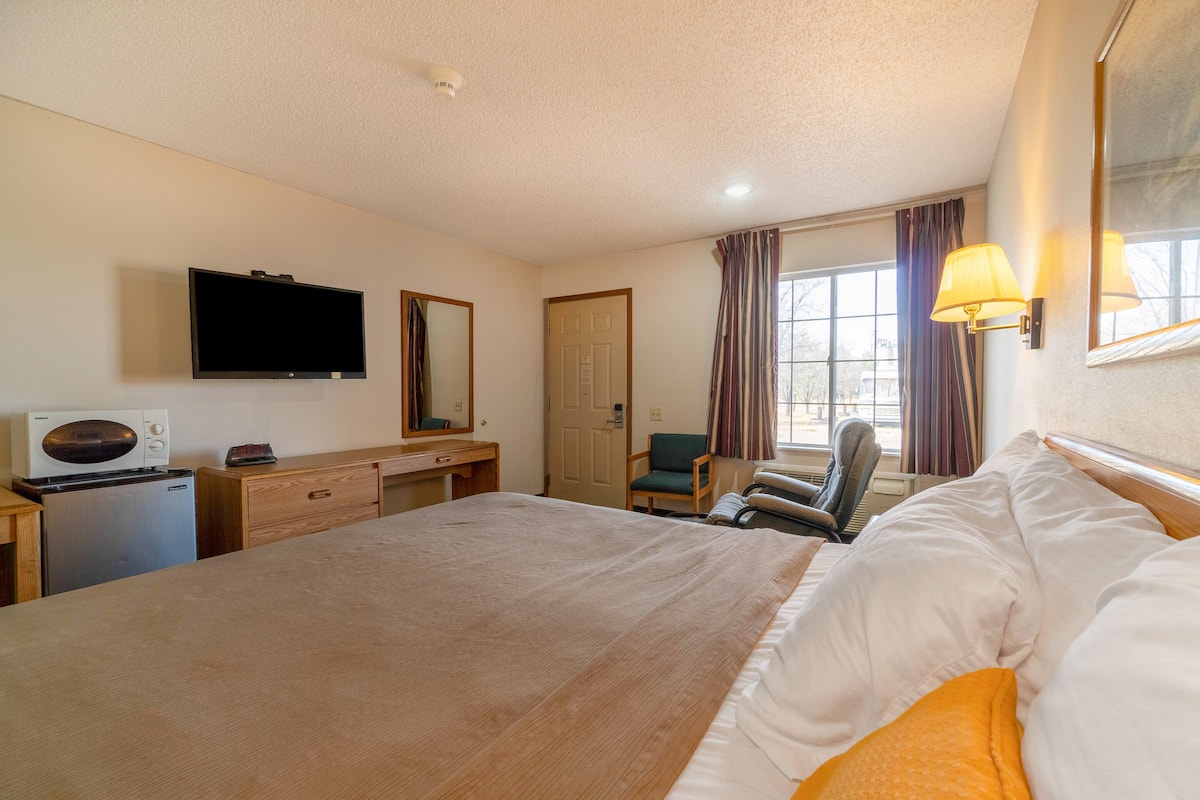 A hotel room with a large bed, a desk with a flat-screen TV, a microwave, a chair, and a window with curtains.