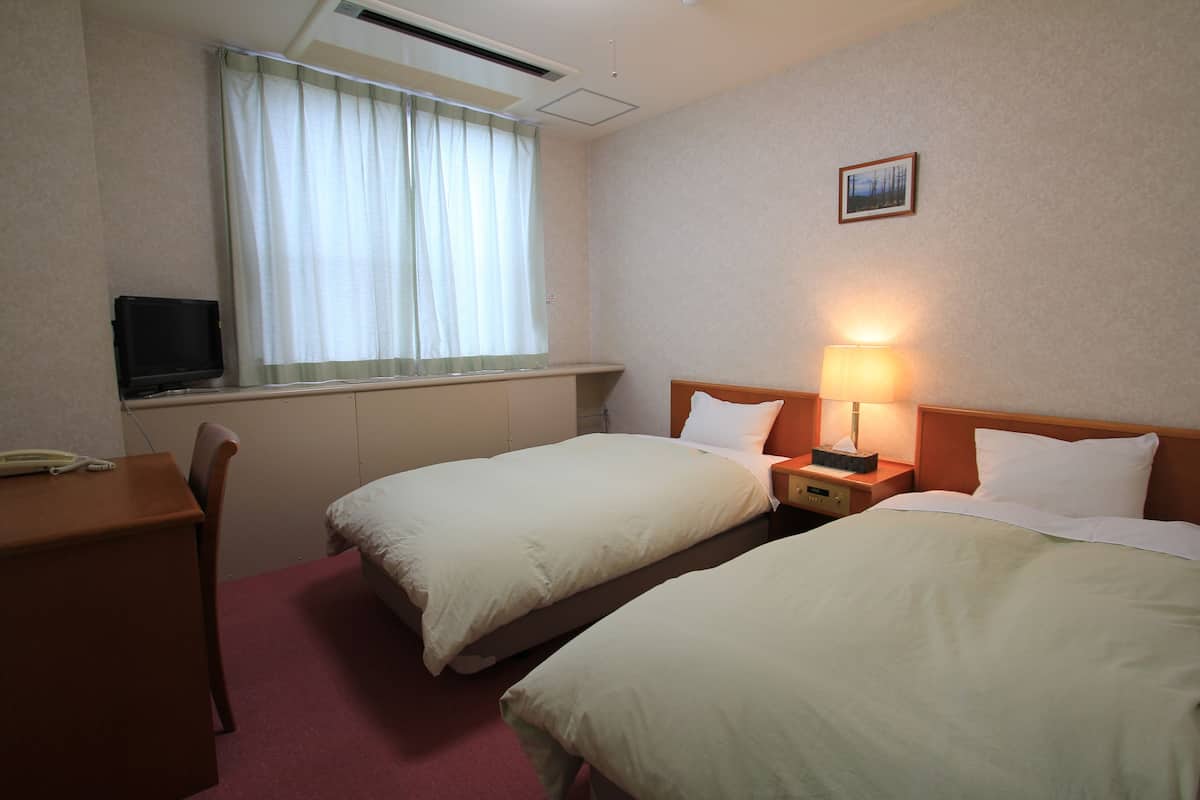 A hotel room with two single beds, a desk with a chair, a television, and a window with curtains.