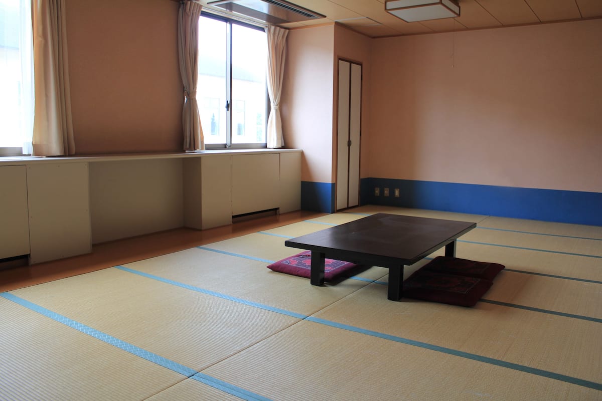A tatami mat room with a low table and cushions, a radiator, and a window with curtains.
