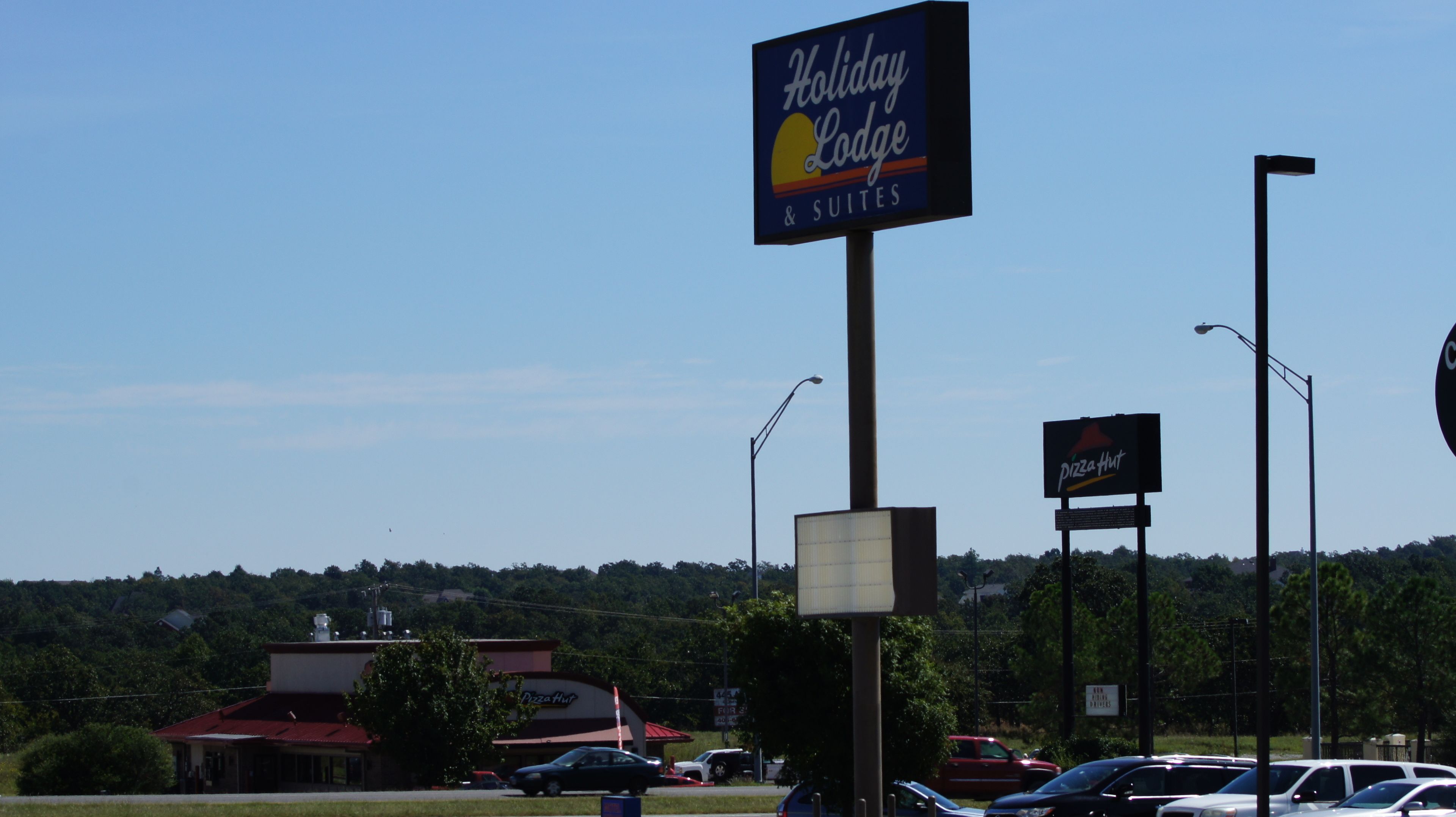 A Holiday Lodge sign with a sunset graphic, a Pizza Hut sign, and a building with a red roof in the background.