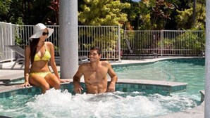 Un hombre y una mujer sentados junto a una piscina con una valla blanca y follaje al fondo.
