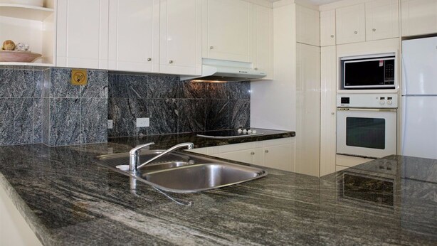 A modern kitchen with granite countertops, stainless steel sink, white cabinets, and built-in appliances including a microwave and oven. - Surfers Mayfair (Surfers Paradise)
