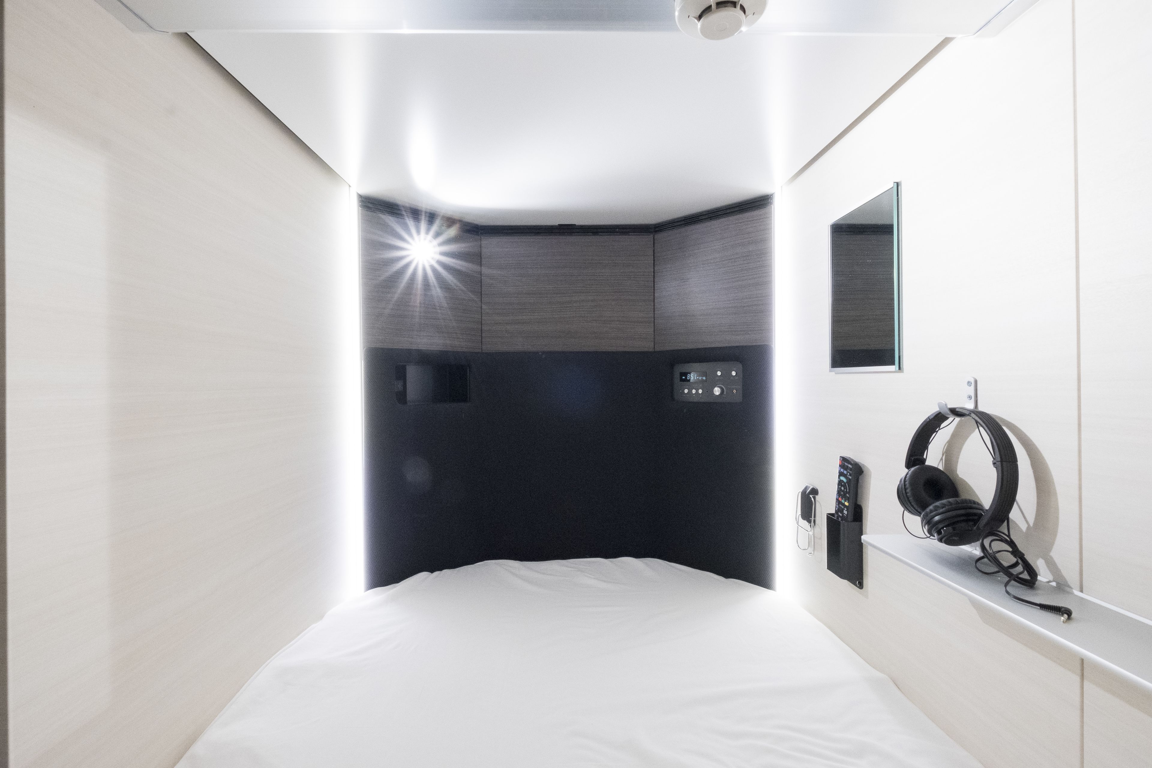 A modern hotel room with a bed, a bedside table with headphones, a phone, and a small shelf.
