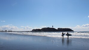 A beach with two people carrying surfboards, a clear sky, and a distant island with a tower. - Fujisawa Hotel En (Fujisawa)