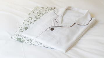A neatly folded white shirt with a brown button and piping on a bed with white sheets and a green leaf-patterned pillow.