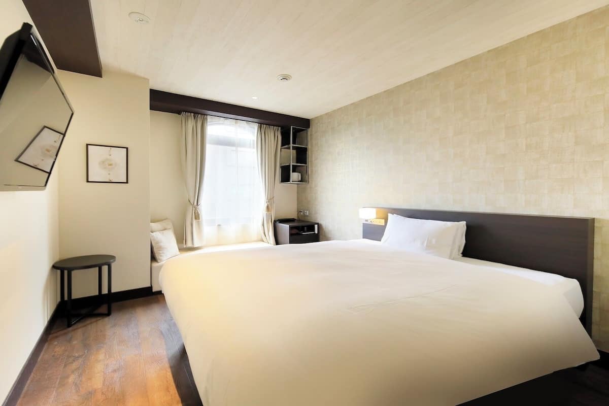 Ein Hotelzimmer mit einem großen Bett, einem Nachttisch, einem Spiegel, einem Fenster mit Vorhängen und einem an der Wand befestigten Regal.