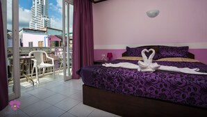 Habitación de hotel con cama, ropa de cama púrpura, una silla blanca y vistas a edificios a través de una puerta de cristal.