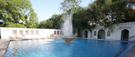 A swimming pool with a central fountain, surrounded by a white building with arched windows and a tiled border.