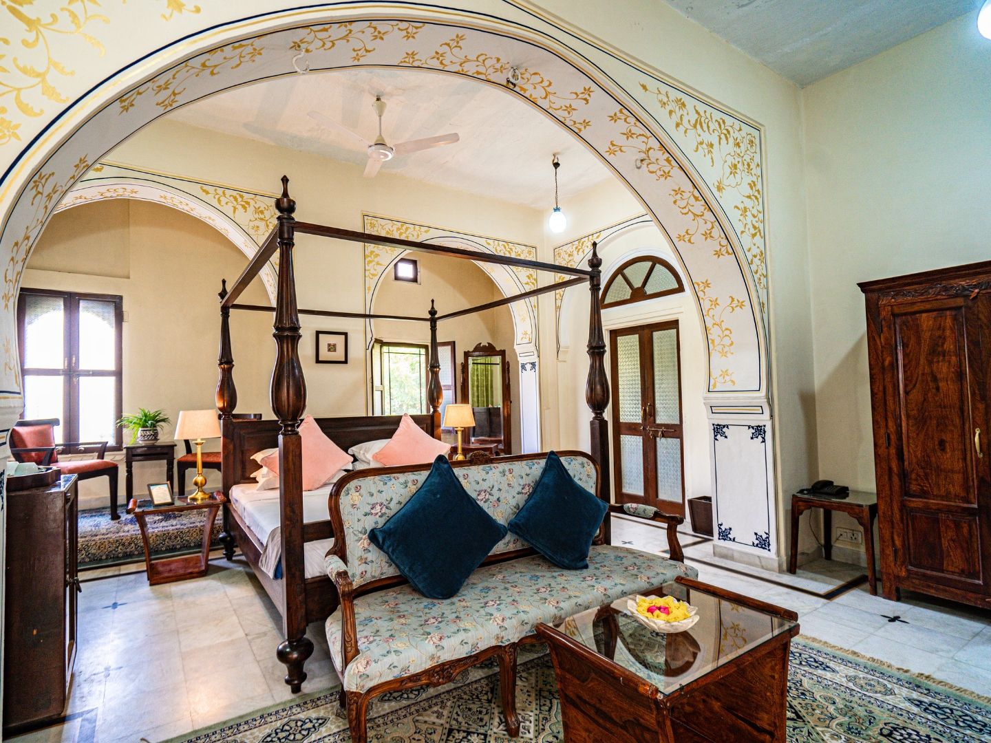 A four-poster bed with a canopy, a sofa with floral upholstery, a wooden coffee table, and a traditional wooden wardrobe.