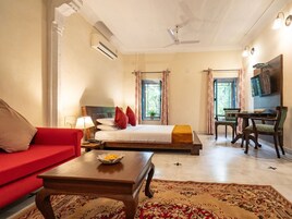 Quarto de hotel com cama, sofá vermelho, mesa de jantar com cadeiras, televisão e ventilador de teto.