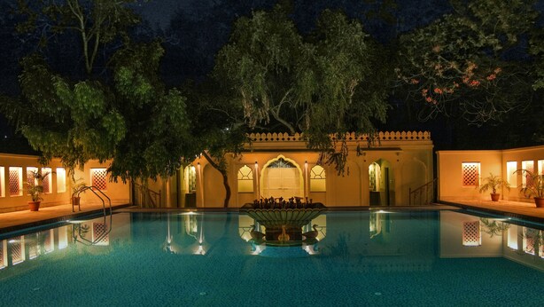 A swimming pool with a fountain, surrounded by trees and a building with arched windows.