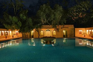 Une piscine avec une fontaine, entourée d’arbres et bordée d’un bâtiment aux fenêtres en arc de cercle.