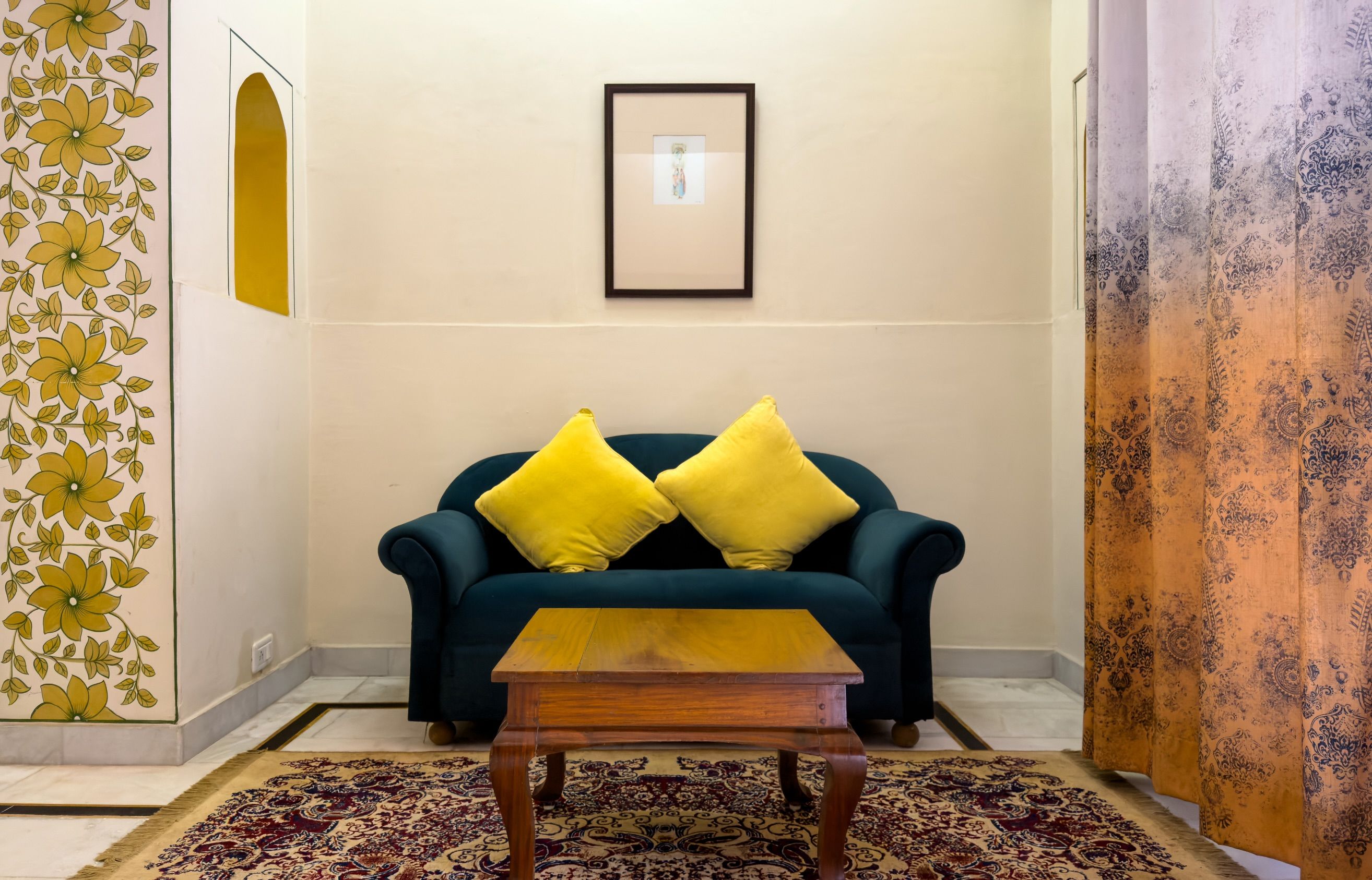 A living room with a dark blue sofa, two yellow cushions, a wooden coffee table, a framed picture on the wall, and patterned curtains.