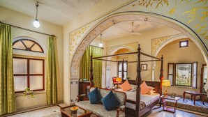 A room with a four-poster bed, a sofa, a small table, and a chair. There is a window with curtains and a ceiling fan.