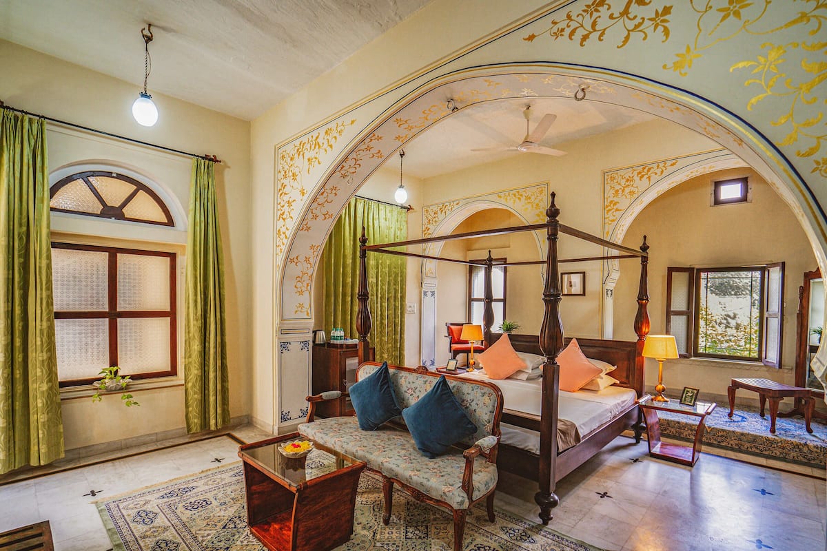 Une chambre avec un lit à baldaquin, un canapé, une petite table et une chaise. Il y a une fenêtre avec des rideaux et un ventilateur de plafond.