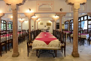 A long dining table with a red and white tablecloth, surrounded by wooden chairs, set in a room with ornate columns and large windows.