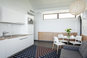 Una cocina moderna con armarios blancos, microondas integrado y un lavamanos. Hay una zona de comedor con una mesa redonda y sillas, y un sofá gris.