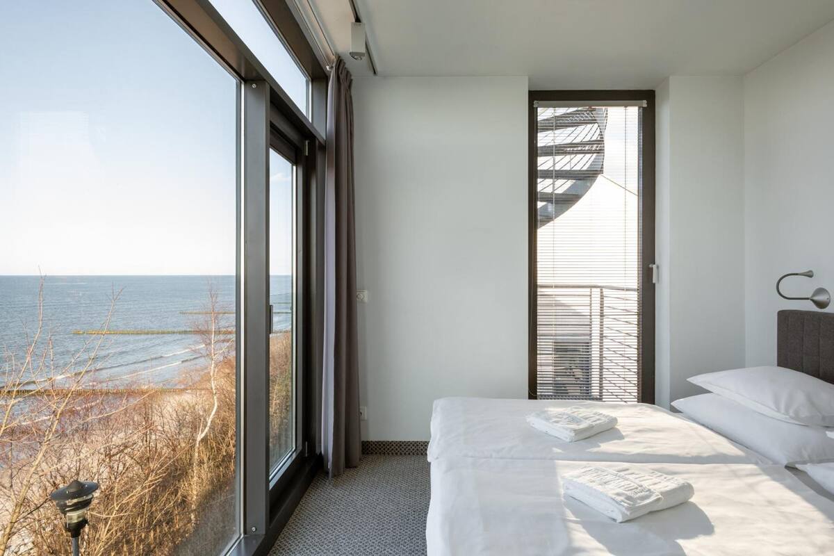 A modern hotel room with a large window offering a view of the sea, a bed with neatly made white linens, and a staircase leading to an outdoor balcony.