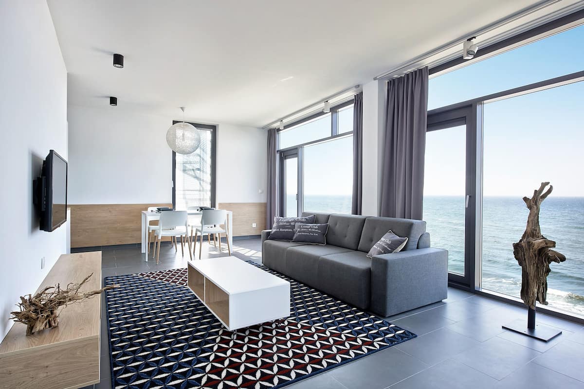 A modern living room with a grey sofa, white coffee table, and patterned rug. There is a dining area with a white table and chairs, and a large window overlooking the sea.