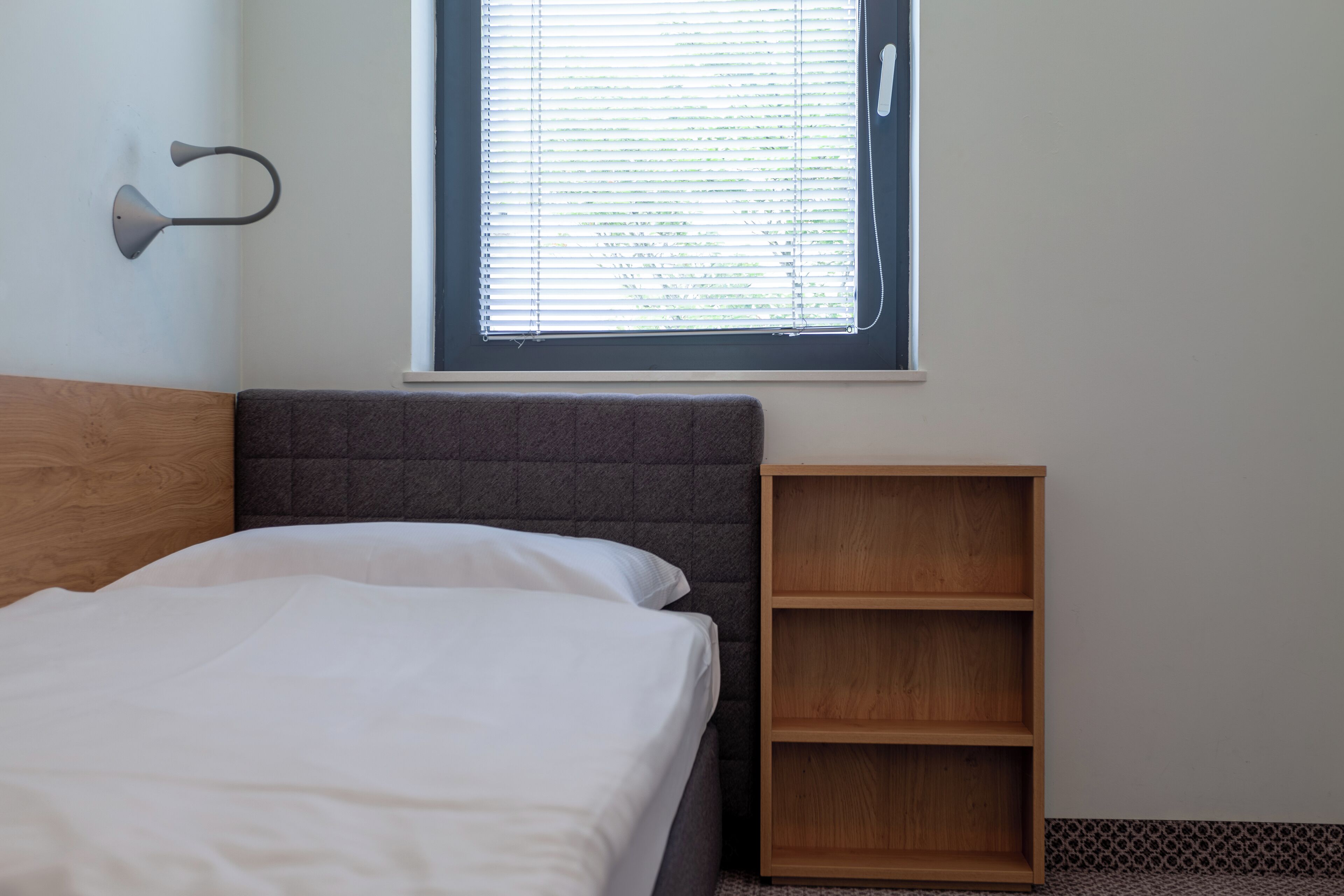 Un dormitorio con cama, mesita de noche, lámpara de pared y ventana con persianas.