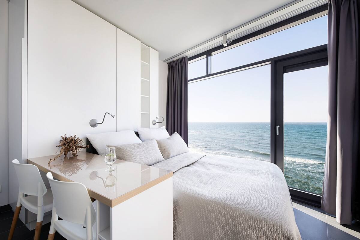 A modern bedroom with a large window overlooking the sea, a wooden desk, and a bed with a white headboard.
