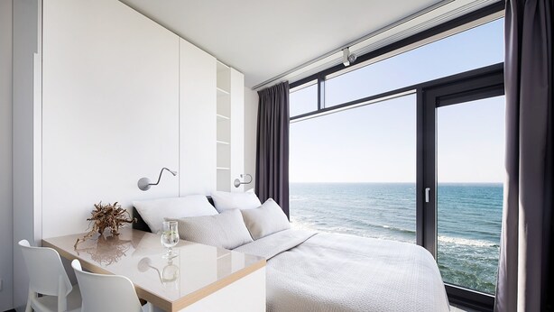 Un dormitorio moderno con un ventanal grande con vistas al mar, un escritorio de madera y una cama con cabecero blanco.