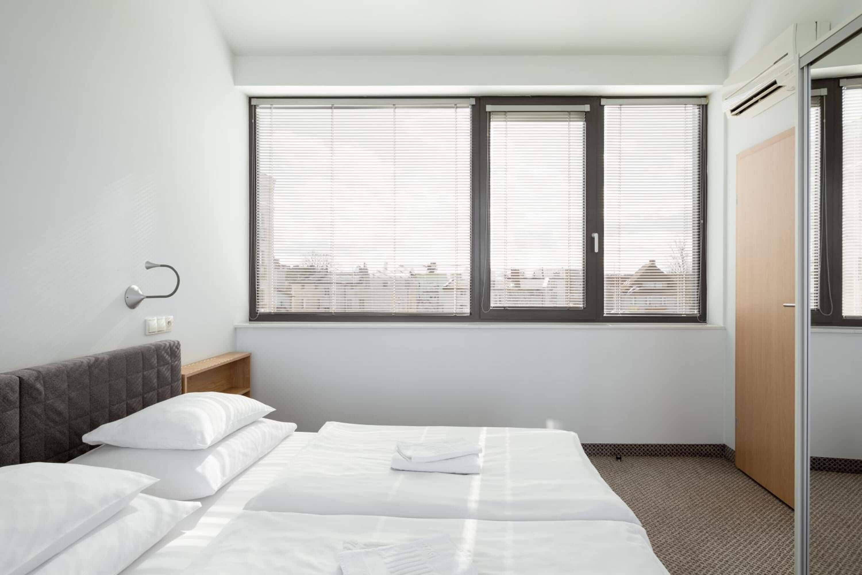 Una habitación de hotel con una cama, dos ventanas, una mesita de noche y un armario.