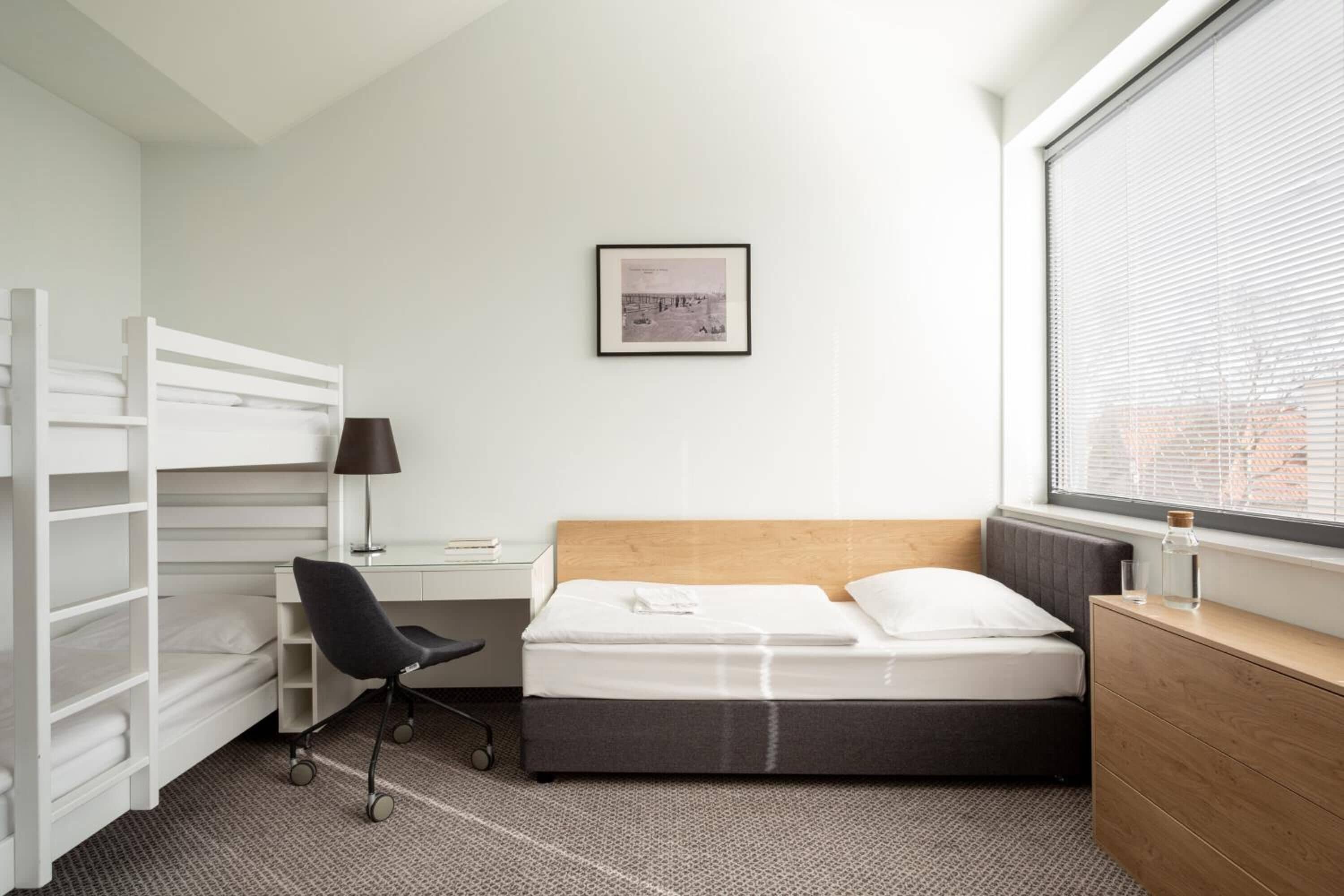 Ein kleines Hotelzimmer mit Etagenbett, Schreibtisch mit Stuhl, Nachttisch und einem Fenster mit Jalousien.