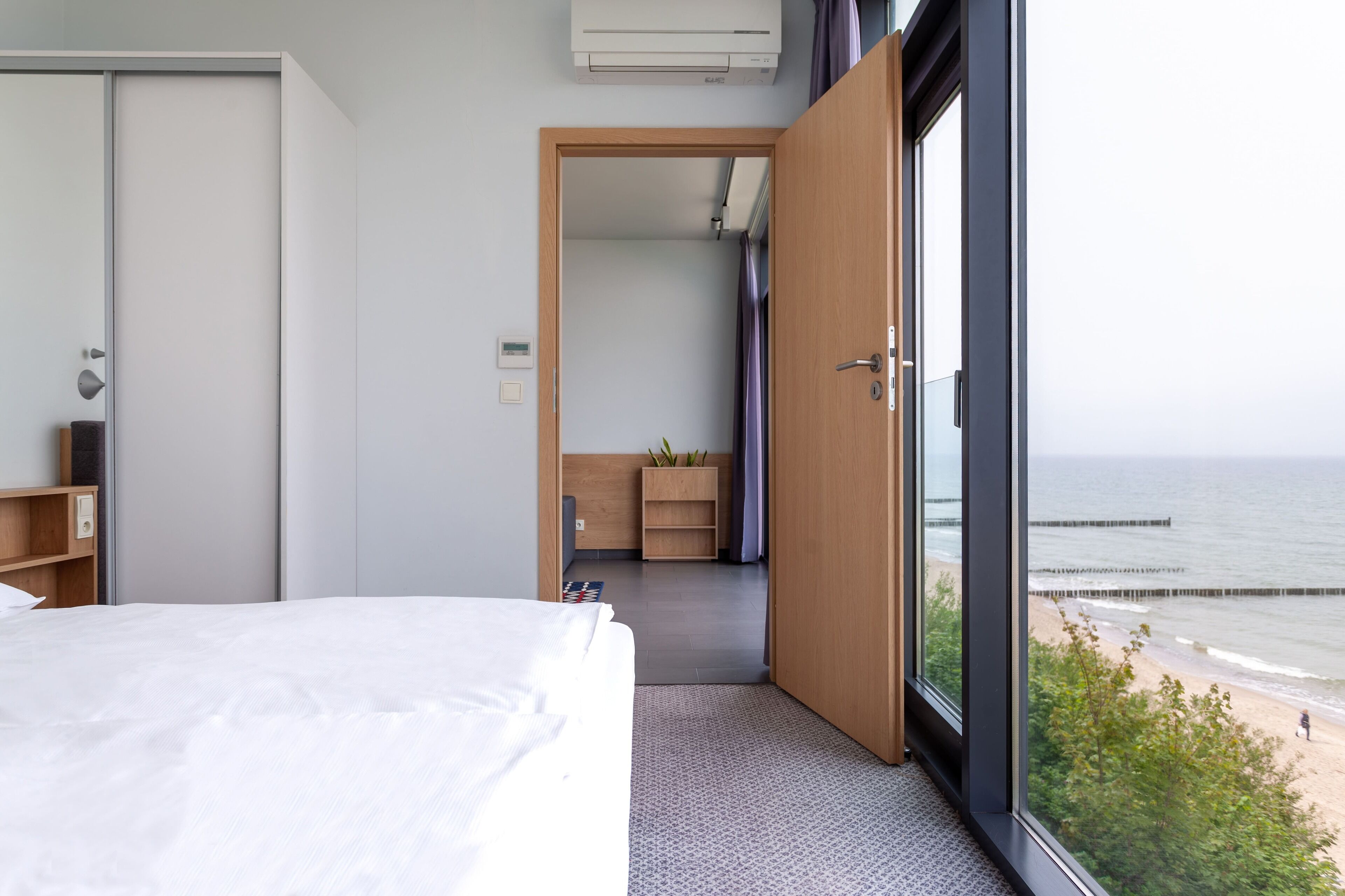 A bedroom with a bed, a wardrobe, a desk, and a view of the ocean through a large window.