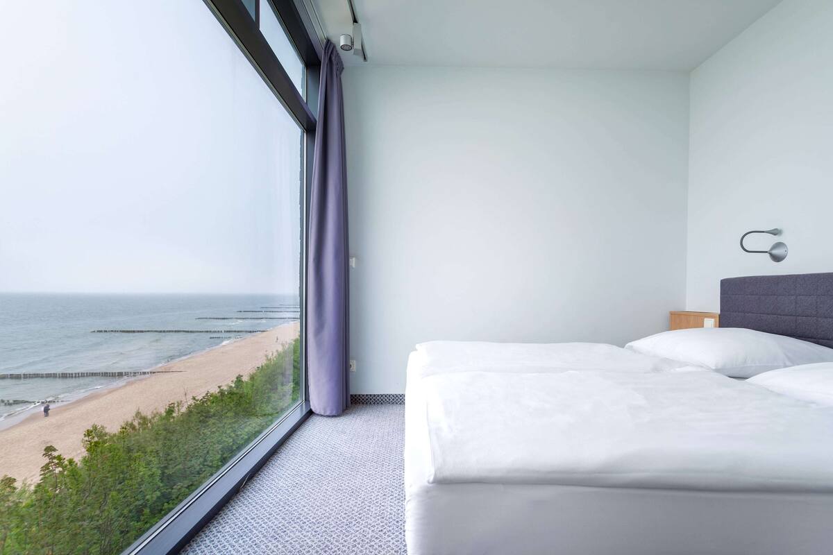 A hotel room with a large window overlooking a beach, a bed with white linens, and a modern headboard.