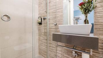 Un baño moderno con ducha envuelta en vidrio, un lavamanos blanco tipo pedestal y un espejo con un florero.