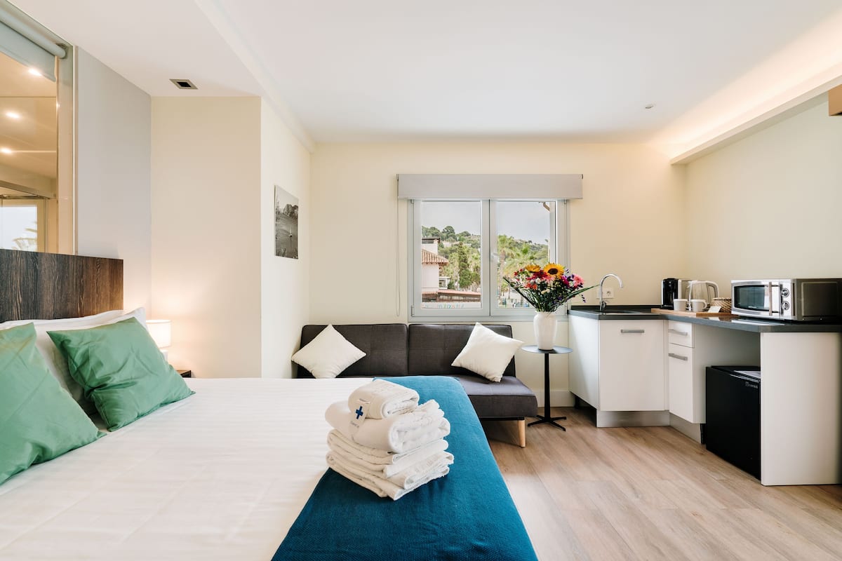 A modern hotel room with a bed, a sofa, a small kitchen area, and a view of the outdoors.