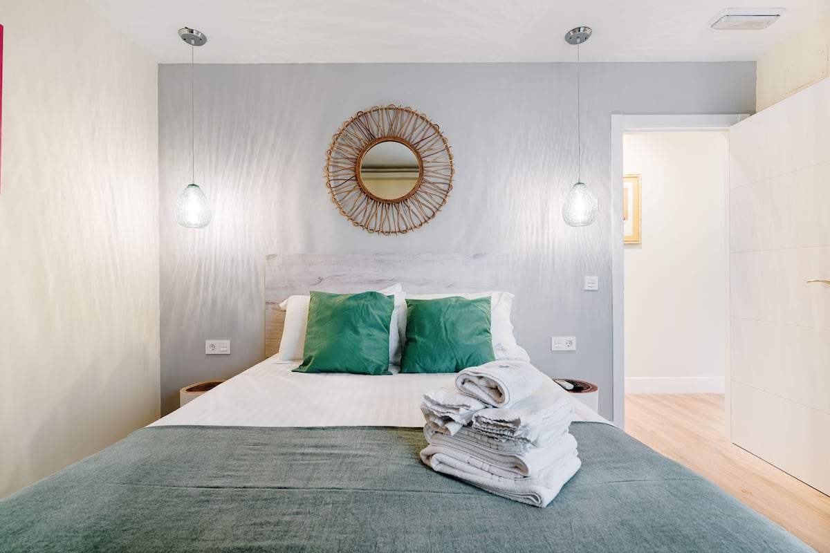 A neatly made bed with green pillows, a round mirror on the wall, and a stack of folded towels on the bed.